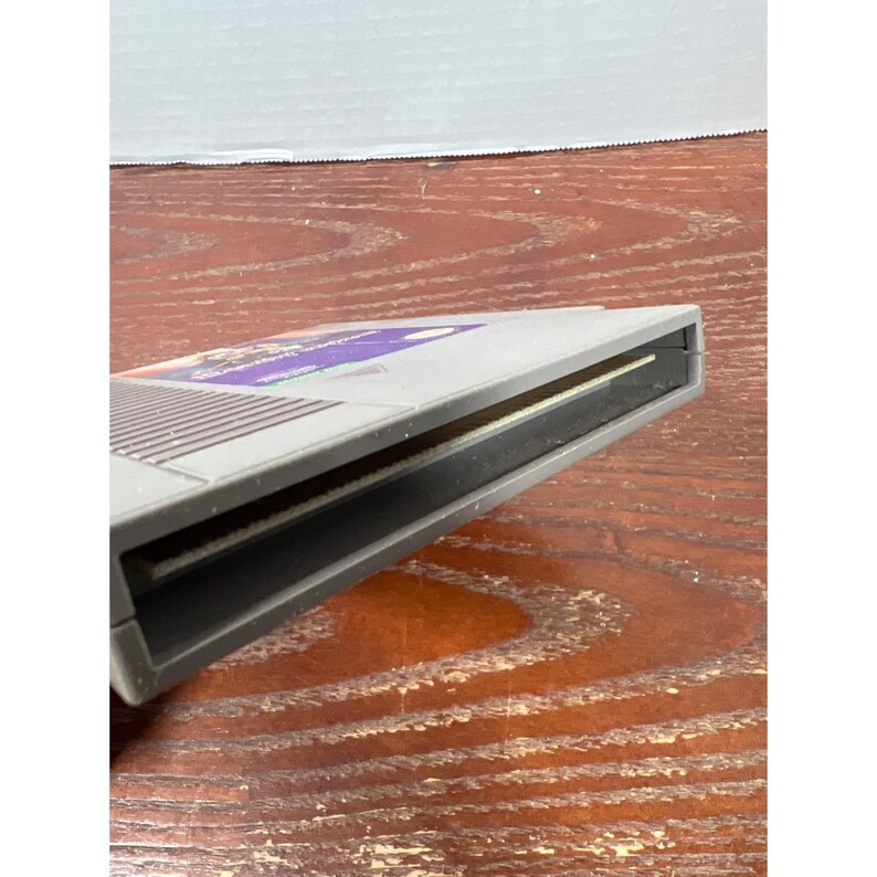 May include: A gray Nintendo Entertainment System (NES) video game cartridge. The cartridge has a purple and white label. The cartridge is rectangular with a slot for insertion into the console. The cartridge is on a wooden surface.