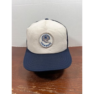 Puede incluir: Una gorra de béisbol con panel frontal blanco y visera y parte trasera azul oscuro. Un parche circular está centrado en la parte delantera, mostrando una estrella azul dentro del contorno de un casco con el texto COWBOYS en azul.