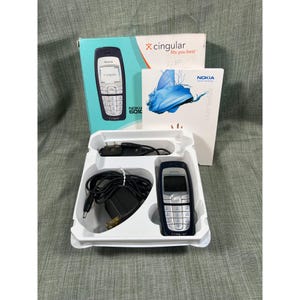Puede incluir: Un teléfono móvil Nokia 6010 vintage en una bandeja de plástico blanco, con un cargador y una guía del usuario. El teléfono es azul oscuro con botones plateados. Una caja con el logotipo de Cingular está en el fondo.
