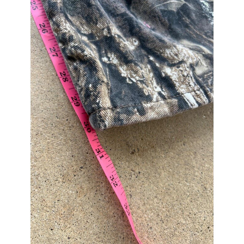 Pode incluir: Close-up de tecido com padr&atilde;o de camuflagem com uma fita m&eacute;trica rosa. A fita mostra medi&ccedil;&otilde;es de 63,5 cm a 81,3 cm. O tecido tem tons de castanho, cinzento e branco.
