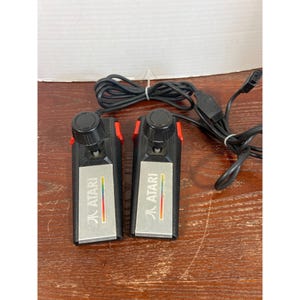 Puede incluir: Dos controladores de paleta Atari negros con una placa frontal plateada que muestra el logotipo de Atari y una franja arcoíris. Cada controlador tiene una perilla circular negra en la parte superior, botones laterales rojos y un cable negro.
