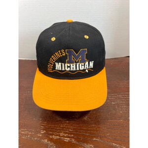 Puede incluir: Una gorra de béisbol negra con visera amarilla y un botón amarillo en la parte superior. El frente presenta texto bordado que dice Wolverines en amarillo, una letra M azul grande con borde amarillo y la palabra Michigan en letras blancas.