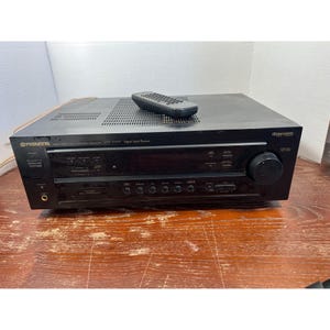 Pode incluir: Um receptor de processador de sinal digital Pioneer VSX-D307 preto com um controle remoto na parte superior. O receptor tem vários botões e um botão de volume. A unidade está em uma superfície de madeira.