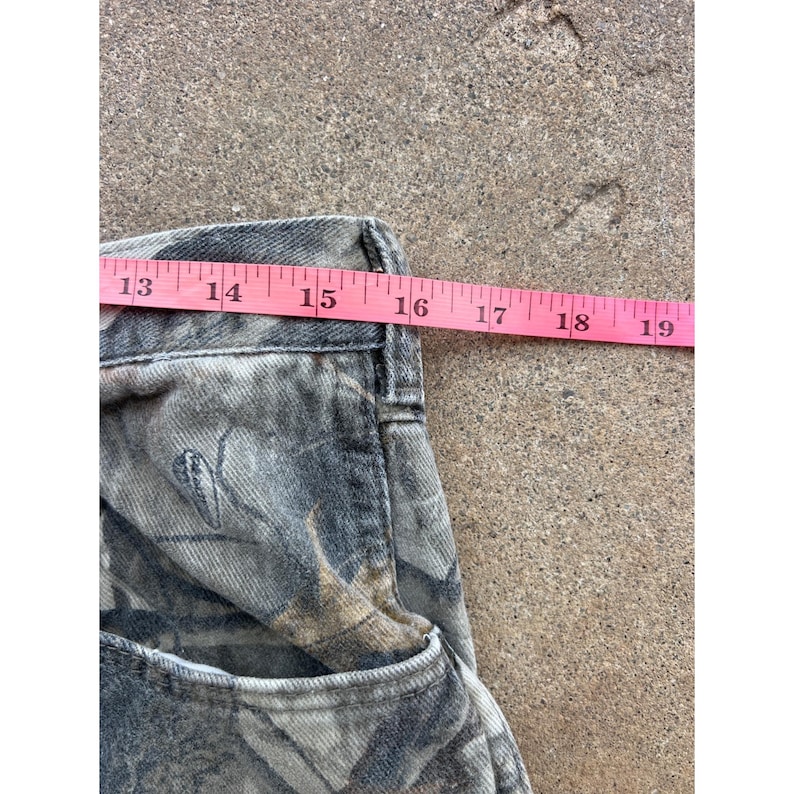 Op de afbeelding: Een broek met camouflageprint in grijs, zwart en bruin. Een roze meetlint is over de tailleband geplaatst en toont een afmeting van ongeveer 48 cm vanaf de zijnaad tot het midden van de taille van het kledingstuk op de afbeelding.