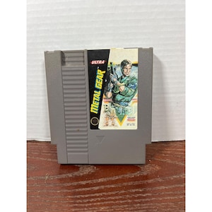 以下が含まれることがあります： 任天堂エンターテインメントシステム用のグレーのメタルギアビデオゲームカートリッジ。ラベルには、銃を持った緑色の軍服を着た男性が描かれています。カートリッジにはUltraのロゴがあります。
