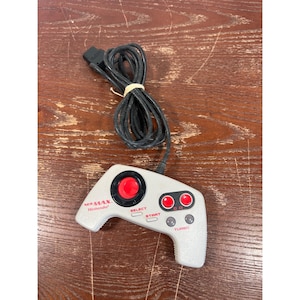 May include: A light gray NES Max video game controller featuring a black circular directional pad with a red center, two red A and B buttons, two gray turbo buttons, and red text reading MS MAX and Nintendo, connected to a black wired cable.