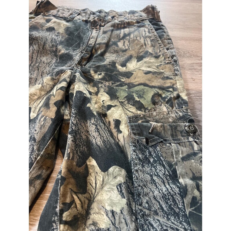 Puede incluir: Pantalones cargo de camuflaje en tonos marrones, verdes y negros. Los pantalones tienen m&uacute;ltiples bolsillos, cierre de bot&oacute;n y cintura con cord&oacute;n. La tela tiene un estampado de hojas.