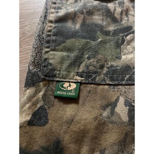 Puede incluir: Primer plano de una tela de camuflaje en tonos verdes, marrones y negros. Una peque&ntilde;a etiqueta verde con las palabras "BRAND CAMO" est&aacute; cosida a la tela. La tela es probablemente parte de una prenda.
