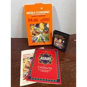 May include: An Atari Missile Command video game set including an orange cardboard box with game artwork, a black plastic game cartridge with a label, and two paper manuals featuring the Atari logo and game catalog information in red and white.