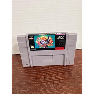 Peut inclure: Une cartouche de jeu vidéo Super Nintendo grise pour le jeu Super James Pond. L'étiquette présente un personnage de dessin animé en costume, le titre, le logo ASC et le sceau de qualité Nintendo. La cartouche a une forme rectangulaire.
