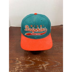 May include: A teal baseball cap with an orange brim and an orange button on top. The front features embroidered text that reads Miami Dolphins Football in orange and white lettering with a script font style and a curved orange underline.