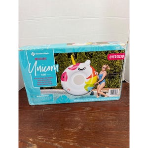 Könnte beinhalten: Ein übergroßer aufblasbarer Einhorn-Schwimmer in einer Box. Der Schwimmer ist weiß mit einem rosa Innenring, einer Regenbogenmähne und einem gelben Horn. Auf der Box steht "Inflatable Unicorn Float" und zeigt eine Frau in einem blauen Badeanzug.