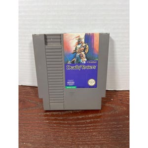 May include: A gray Nintendo Entertainment System (NES) game cartridge for the game "Deadly Towers." The label features a warrior illustration with the game title in a blue box. The cartridge is rectangular and made of plastic.