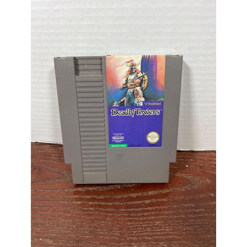 May include: A gray Nintendo Entertainment System (NES) game cartridge for the game "Deadly Towers." The label features a warrior illustration with the game title in a blue box. The cartridge is rectangular and made of plastic.