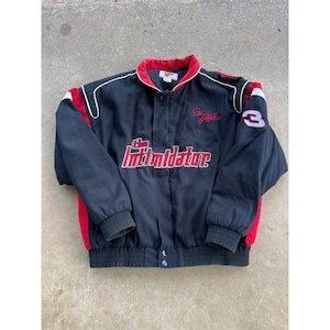 Puede incluir: Chaqueta negra con detalles rojos y blancos. Presenta el texto "The Intimidator" en rojo y blanco, junto con el número "3" en la manga. La chaqueta tiene cuello y puños rojos, y cierre de botones.