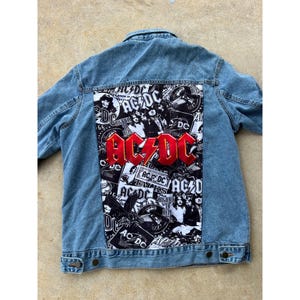 May include: Pale blue denim jacket featuring a large AC/DC graphic on the back. The graphic includes the band's red logo and black and white images of the band. The jacket has a classic button-up design.