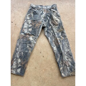 Op de afbeelding: Broek van volledige lengte met een realistisch boscamouflagepatroon in tinten bruin, grijs en zwart. Deze broek heeft een knoopsluiting, twee voorzakken en verstevigde kniestukken, wat zorgt voor een gestructureerde pasvorm met rechte pijpen.