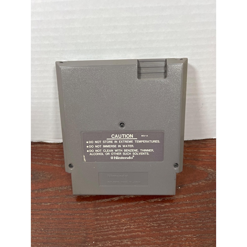 May include: A gray Nintendo game cartridge with a rectangular shape. The back of the cartridge has a label with the word "CAUTION" and instructions. The Nintendo logo is visible at the bottom of the label.