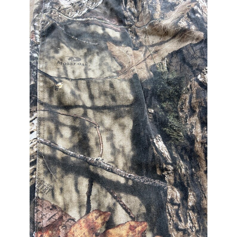 Pode incluir: Close-up de tecido camuflado com um padr&atilde;o marrom, verde e bege. O tecido tem uma apar&ecirc;ncia texturizada e as palavras "Mossy Oak" s&atilde;o vis&iacute;veis.