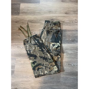 May include: Camouflage cargo pants in shades of green, brown, and black. The pants have multiple pockets and a drawstring waist. The fabric has a woodland pattern, suitable for outdoor activities.