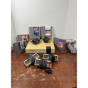 May include: A vintage Nintendo Entertainment System (NES) console with several game cartridges, controllers, and a light gun. The console is beige and black, with multiple game cartridges featuring colorful artwork. The controllers are gray with red and black buttons.