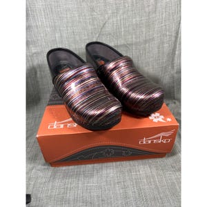 May include: A pair of Dansko XP clogs featuring a glossy, multi-colored striped pattern in shades of purple, blue, and metallic silver. The shoes are positioned on top of an orange Dansko branded shoebox with white text and logo details.