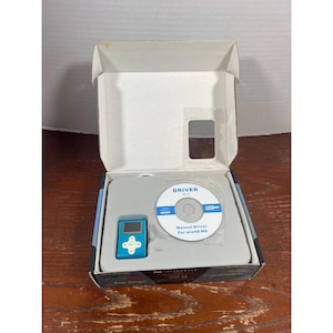 May include: A blue MP3 player with a small screen and control buttons, packaged in a box with a CD-ROM labeled "DRIVER A-1". The box also contains a manual for Win98/ME. The product is displayed on a wooden surface.