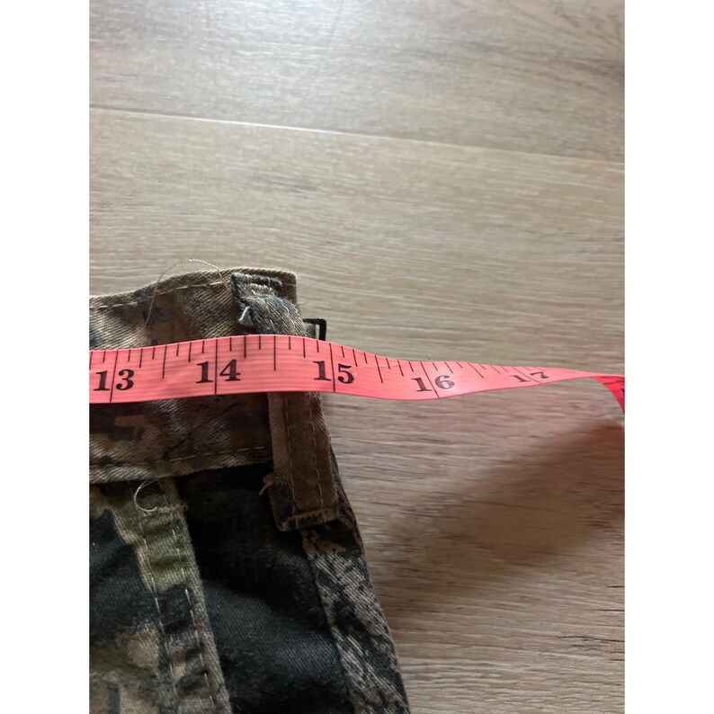 Puede incluir: Primer plano de una tela con estampado de camuflaje con una cinta m&eacute;trica rosa estirada sobre ella. La cinta lee de 33 a 40 cm. La tela parece ser parte de una prenda, posiblemente pantalones cortos o largos.