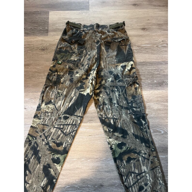 Puede incluir: Pantalones cargo de camuflaje en tonos marrones, verdes y negros. Los pantalones tienen m&uacute;ltiples bolsillos, un cintur&oacute;n y un cierre de bot&oacute;n. La tela tiene un aspecto texturizado, y los pantalones est&aacute;n planos sobre una superficie de madera.