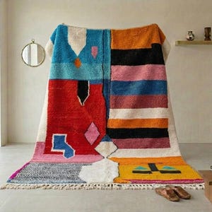 May include: A colorful, geometric patterned rug with a mix of blue, red, orange, pink, and black sections. The rug has a fringed edge and is displayed indoors, leaning against a wall. The design includes various shapes and patterns.
