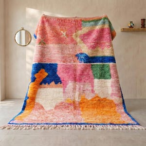 Op de afbeelding: Een groot, rechthoekig, handgeknoopt wollen tapijt met een levendig, abstract geometrisch patroon in tinten roze, oranje, blauw, groen en crème. Het tapijt heeft een dikke, hoogpolige textuur en is aan de onderkant afgewerkt met crèmekleurige franjes.