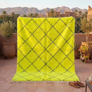 Puede incluir: Una alfombra amarilla brillante con un patrón de diamantes negros. La alfombra se exhibe al aire libre, con una vista de montañas y palmeras al fondo. La alfombra tiene una textura peluda y una franja en el borde inferior.