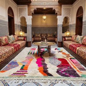 Hand-knotted plush Berber wool shag rug in natural ivory cream, featuring free-form vertical abstract color columns and figure forms in fuchsia pink, teal, orange, crimson, forest green, speckled gray-blue, blush, mauve, purple, and amber rising the full height of the field, with a pale sage green lower border stripe and natural cream fringe, photographed flat on a sunlit natural stone surface in Morocco.