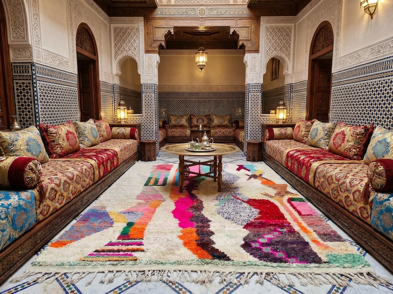 Hand-knotted plush Berber wool shag rug in natural ivory cream, featuring free-form vertical abstract color columns and figure forms in fuchsia pink, teal, orange, crimson, forest green, speckled gray-blue, blush, mauve, purple, and amber rising the full height of the field, with a pale sage green lower border stripe and natural cream fringe, photographed flat on a sunlit natural stone surface in Morocco.