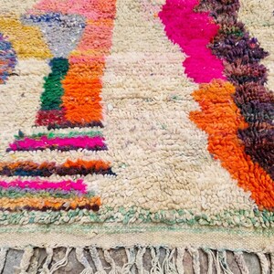May include: A thick, hand-knotted wool rug featuring a cream-colored base with an abstract pattern of vibrant, multi-colored shapes in shades of hot pink, bright orange, deep purple, green, and blue, finished with a knotted fringe along the edge.