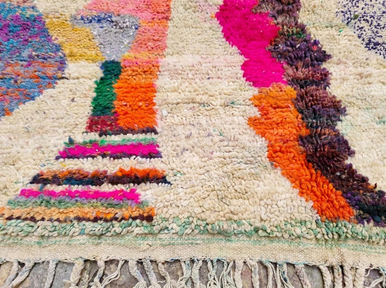 May include: A thick, hand-knotted wool rug featuring a cream-colored base with an abstract pattern of vibrant, multi-colored shapes in shades of hot pink, bright orange, deep purple, green, and blue, finished with a knotted fringe along the edge.