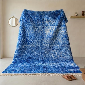 Puede incluir: Una alfombra grande, azul y blanca texturizada con un borde con flecos. La alfombra se exhibe en interiores, sobre una estructura, con un fondo neutro. El diseño de la alfombra tiene una apariencia moteada, con tonos de azul y blanco.