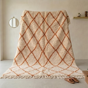 May include: A large, cream-colored rug with a repeating diamond pattern in a warm orange hue. The rug has a thick, textured pile and a fringe along the bottom edge. The rug is displayed indoors, leaning against a wall.