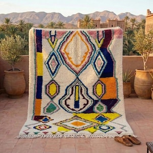 May include: A vibrant, geometric patterned rug with a cream backdrop. The rug showcases diamond and oval shapes in yellow, blue, orange, and pink. The rug is displayed outdoors on a terracotta surface, with plants and a mountain range in the background.