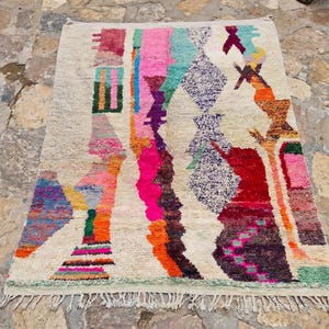 May include: A rectangular area rug with a cream-colored base featuring an abstract, multi-colored pattern. The design includes vertical bands and geometric shapes in vibrant shades of pink, orange, purple, teal, red, and green with fringe ends.