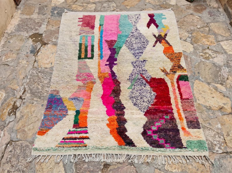 May include: A rectangular area rug with a cream-colored base featuring an abstract, multi-colored pattern. The design includes vertical bands and geometric shapes in vibrant shades of pink, orange, purple, teal, red, and green with fringe ends.