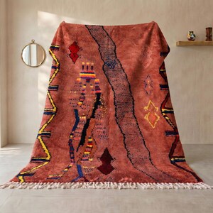 May include: A large, rectangular, terracotta-coloured rug featuring a thick, dark, textured vertical stripe and various geometric patterns in yellow, blue, and dark red. The rug has a deep pile and a fringe of white tassels along the bottom edge.
