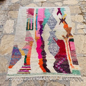 May include: A rectangular area rug with a cream-colored base featuring a vibrant, abstract pattern of vertical stripes and geometric shapes in shades of hot pink, turquoise, orange, purple, red, and yellow, finished with a knotted fringe at the bottom.