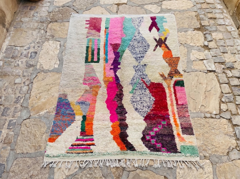 May include: A rectangular area rug with a cream-colored base featuring a vibrant, abstract pattern of vertical stripes and geometric shapes in shades of hot pink, turquoise, orange, purple, red, and yellow, finished with a knotted fringe at the bottom.