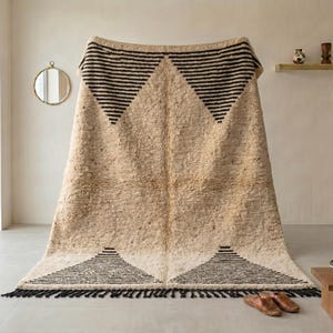 May include: A beige and black geometric patterned rug with a textured surface. The rug features black triangle designs and a fringe along the bottom edge. The rug is displayed indoors.