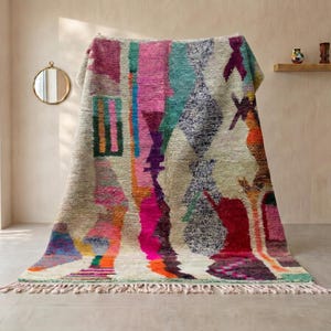 May include: A large rectangular area rug featuring a cream-colored base with an abstract pattern of vertical stripes and geometric shapes in vibrant shades of hot pink, teal, orange, purple, and dark gray, finished with a knotted fringe at the bottom.