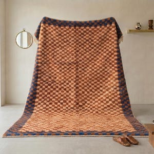 May include: A large, rectangular woven blanket with a brown and blue checkered pattern. The blanket is displayed indoors, with a neutral background and natural light. The edges are trimmed with a dark blue border and fringe.