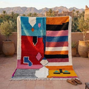May include: A colorful, geometric patterned rug with a white border. The rug features a mix of blue, red, orange, black, pink, and yellow sections. The rug is displayed outdoors, with a view of mountains and palm trees in the background.