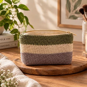 Puede incluir: Un estuche rectangular de ganchillo con tres franjas horizontales de colores en verde oliva, crema y morado apagado. El accesorio está confeccionado con un patrón de punto denso y texturizado e incluye una cremallera metálica dorada.