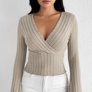 Peut inclure: Haut beige en tricot côtelé avec un col en V croisé et des manches longues. Les manches s'évasent aux poignets. Le haut est associé à un pantalon blanc. Le modèle a les cheveux noirs et regarde la caméra.